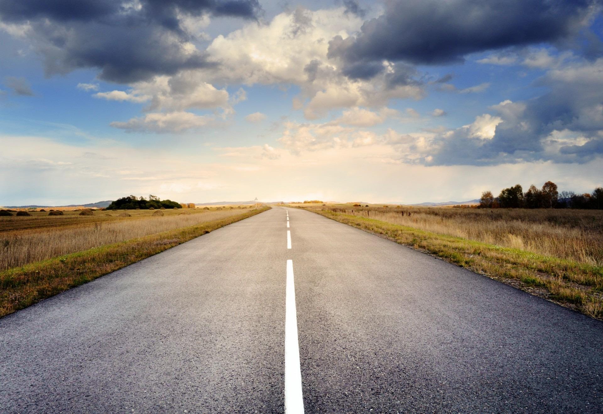 Straight asphalt road in Nebraska under a partly cloudy sky, bordered by grassy fields and distant trees.