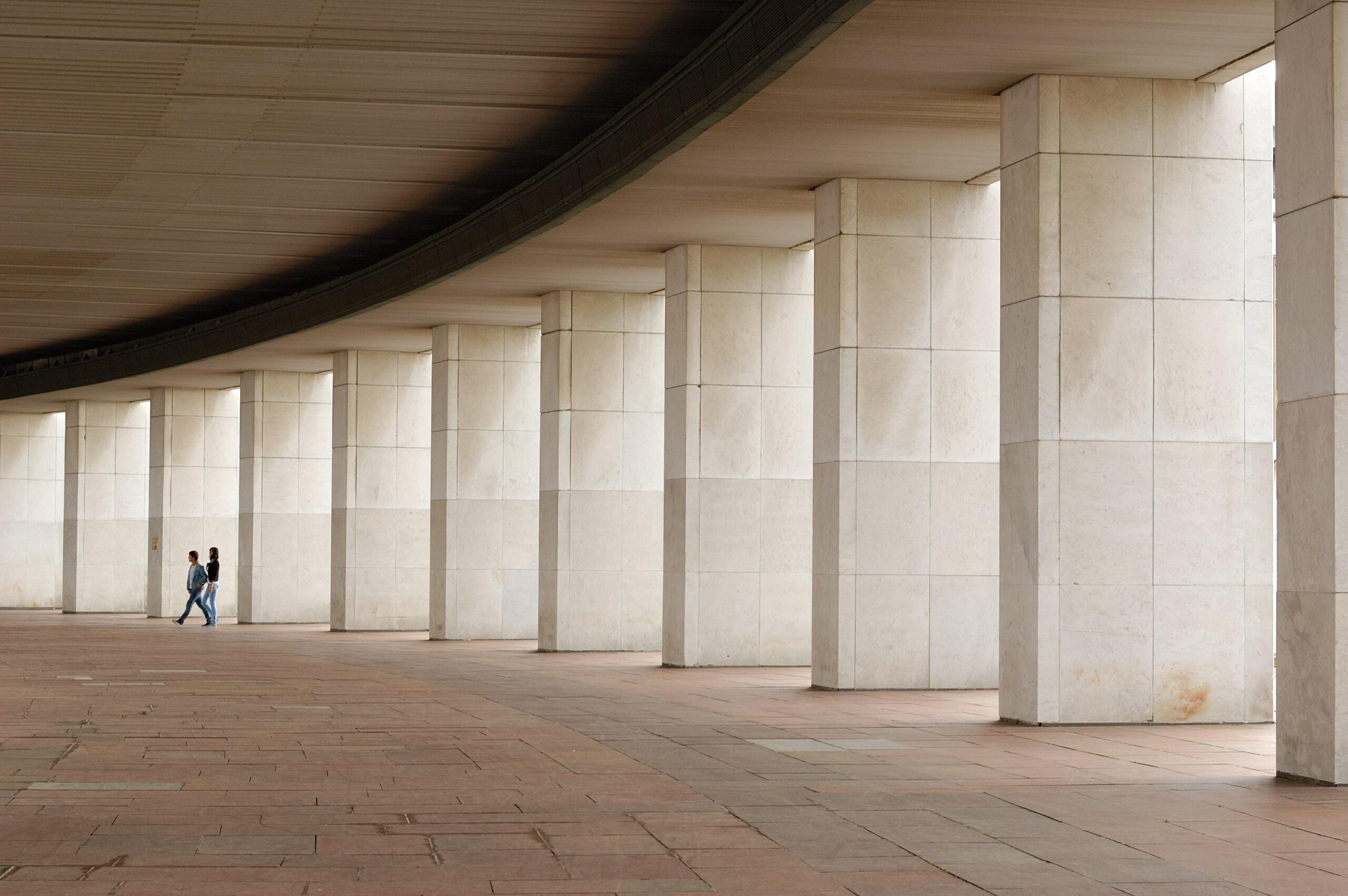 Two people walking between concrete architectural columns at a construction site, illustrating prefab concrete panels and modular construction logistics.